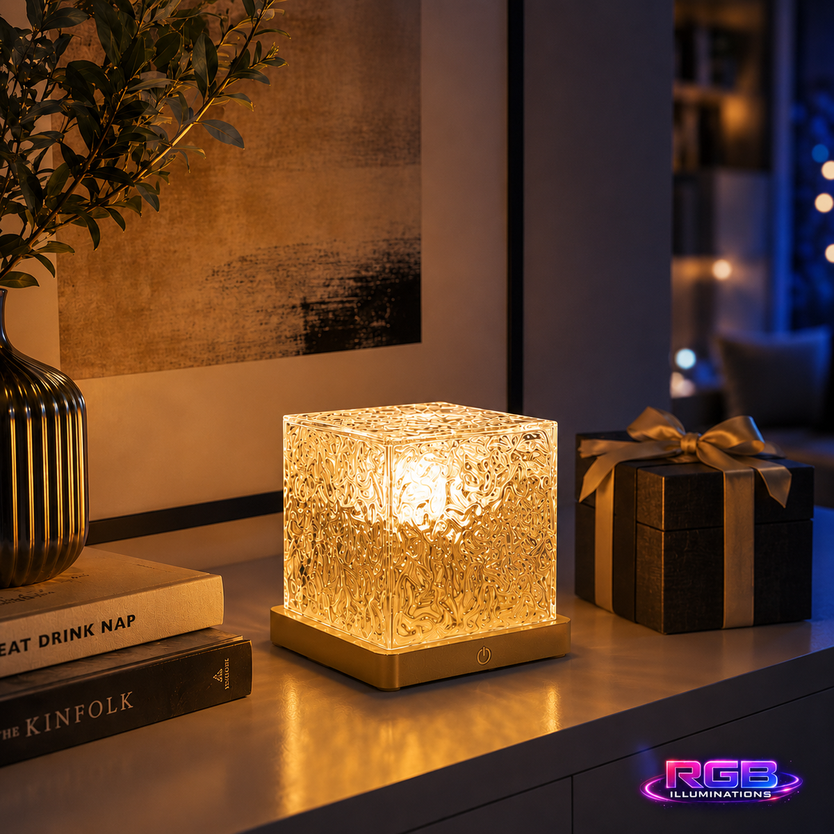 A glowing square LED night light with a textured, crackle-glass-like front and a warm gold base, styled on a tabletop next to books, a vase, and a gift box in a dimly lit room. — RGB Illuminations product view 4