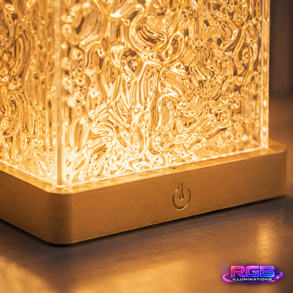 A close-up of a glowing square lamp with an intricate, textured glass or acrylic front panel and a metallic gold base. The lamp is turned on, casting a warm golden light onto a dark surface.