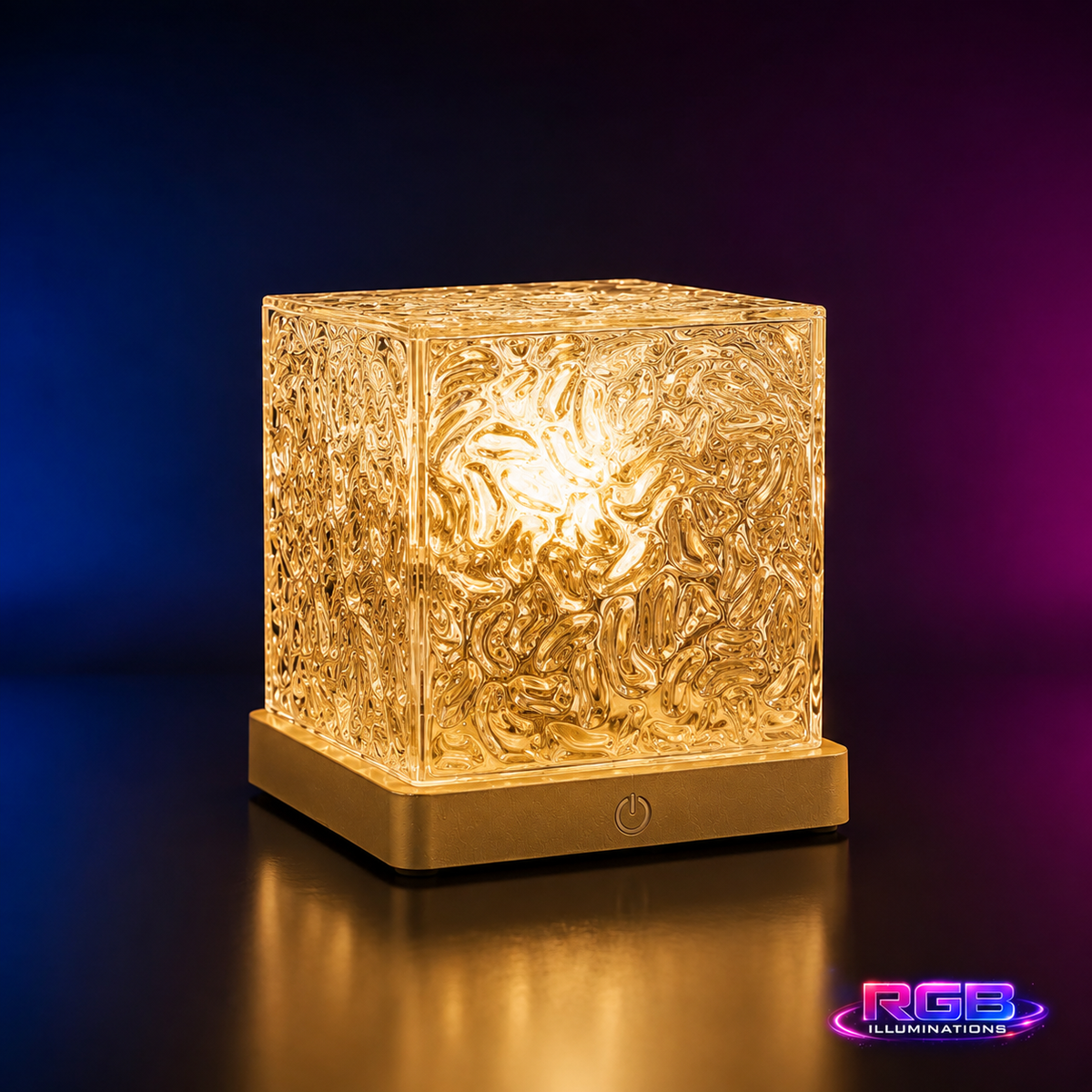 A cube-shaped LED light fixture with a textured, crackled clear acrylic front panel that glows warm yellow when illuminated, sitting on a square metallic gold base. The background is a dark studio setting with blue and purple gradient lighting, and an 'RGB ILLUMINATIONS' logo is visible in the bottom right corner. — RGB Illuminations product view 11