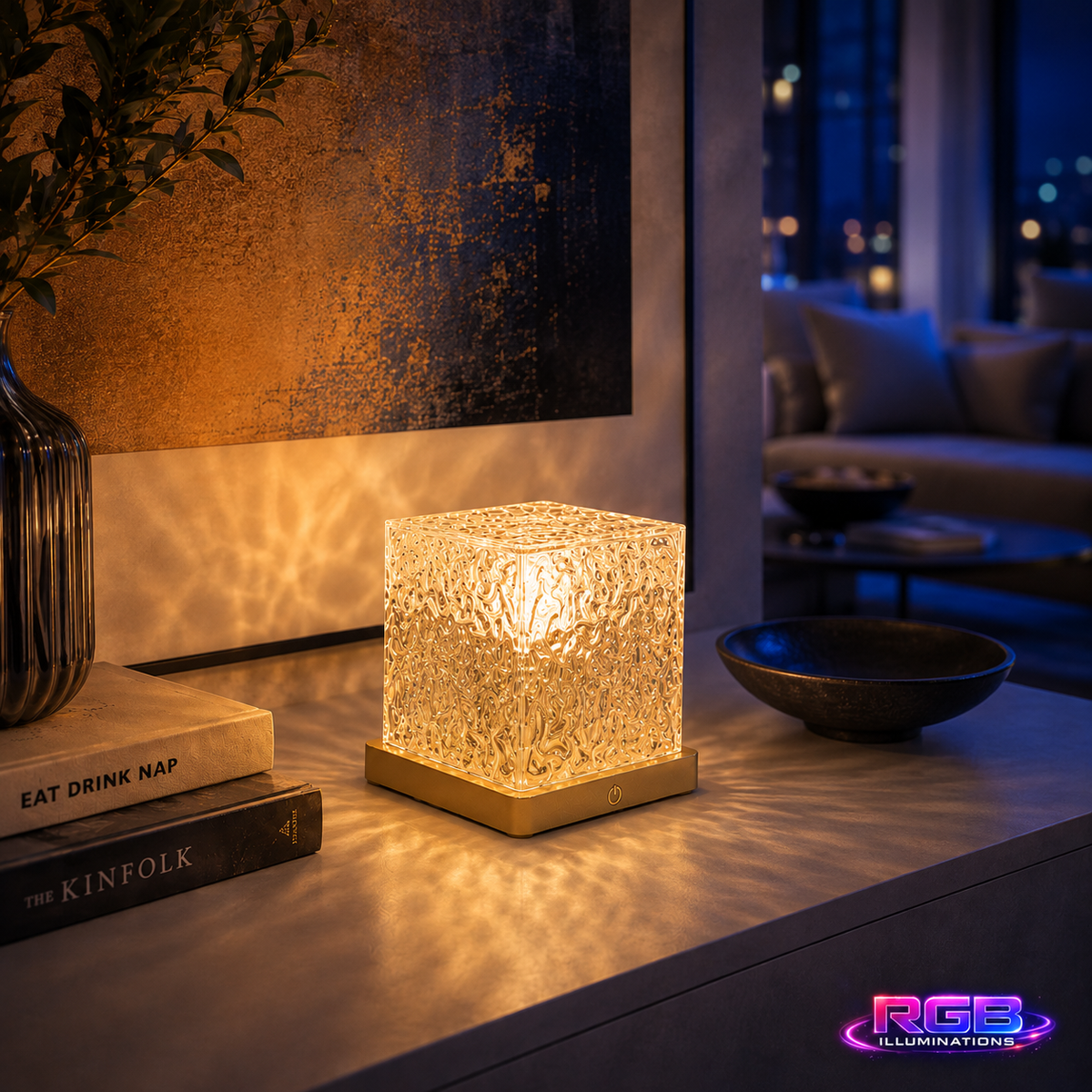 A glowing square LED table lamp with a textured, crackle-glass-like cube body and a gold-toned base, placed on a tabletop in a modern living room setting. — RGB Illuminations product image