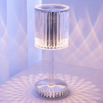 A sleek, minimalist table lamp with a cylindrical, colour-changing LED design, mounted on a thin, metallic stand, with a remote control.