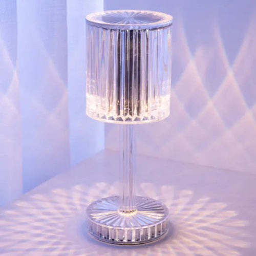 A sleek, minimalist table lamp with a cylindrical, colour-changing LED design, mounted on a thin, metallic stand, with a remote control. RGB Illuminations